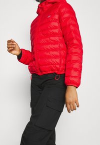 Chaqueta acolchada ligera roja con capucha, diseño acolchado horizontal, dos bolsillos laterales y puños elásticos. Combinada con pantalones cargo negros.