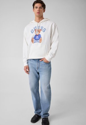 Junger Mann trägt weißen Hoodie mit Teddybär und "GUESS"-Schriftzug, hellblaue Jeans und schwarze Mokassins vor einfarbigem Hintergrund.