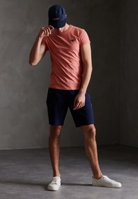 Röd texturerad t-shirt med korta ärmar och logotyp, ihop med marinblå shorts och vita sneakers. Kappen har en mörkblå färg med en logotyp.