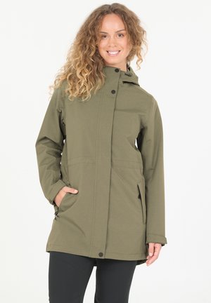 Femme aux cheveux bouclés portant une veste de pluie verte à capuche avec des poches zippées et un pantalon noir, souriant avec une main dans la poche.