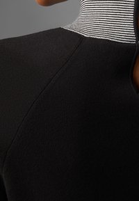 Svart tyg med en texturerad yta, med en hög krage i omväxlande svarta och vita ränder. Dragkedjestängning bak.