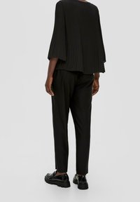 Svart plisserad topp med breda ärmar, matchad med svarta smala byxor och kraftiga svarta skor, sedd bakifrån.