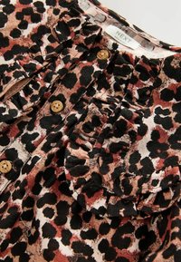 Blouse à imprimé léopard avec un col froncé, taches noires et brunes sur fond crème, dotée de boutons en bois et d'un tissu texturé.