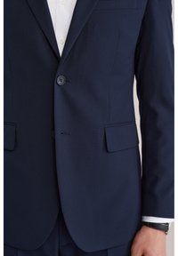 Granatowa marynarka garniturowa zapinana na jeden guzik, na białej koszuli, pokazująca dolną część tułowia i prawy ramię z czarnym zegarkiem na nadgarstku.