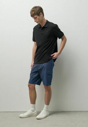 Junger Mann, der ein schwarzes Poloshirt, blaue Cargoshorts, weiße Socken und weiße Turnschuhe trägt, vor einer schlichten hellgrauen Wand stehend.