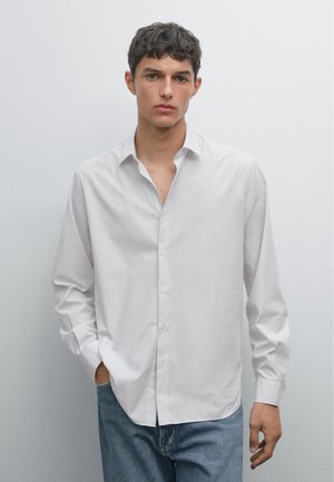 Witte gestreepte button-up shirt met een ontspannen pasvorm en kraag, gecombineerd met blauwe denim jeans. Lange mouwen, zachte textuur en subtiele strepen.