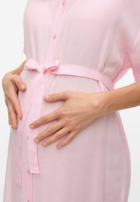 Robe boutonnee rose clair en tissu transparent, dotée d'une ceinture nouée à la taille. Des mains reposent doucement sur un ventre arrondi.