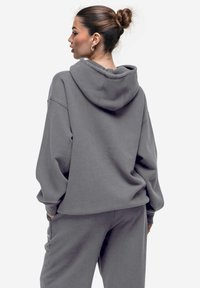 Hoodie grigio oversize in materiale morbido con cappuccio regolabile, spalle cadenti e vestibilità rilassata, abbinato a pantaloni coordinati.