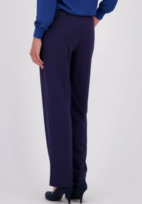 Vrouw in donkerblauwe rechte broek, bijpassend blauwe top en donkerblauwe hoge hakken, staand tegen een neutrale achtergrond.