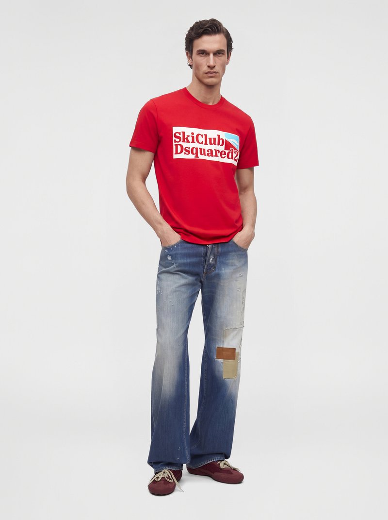 Мъжки модел стои с ръце в джобовете, облечен с червена тениска SkiClub Dsquared2, широки избелели дънки и бордо маратонки.