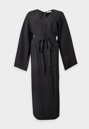 Femme aux longs cheveux foncés portant une robe portefeuille noire à larges manches, nouée à la taille, et des chaussures à talons beiges sur un fond uni.