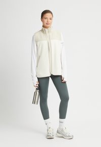 Kvinna i beige väst, vit långärmad skjorta, grå leggings, vita strumpor och sneakers som håller en vattenflaska i rostfritt stål.