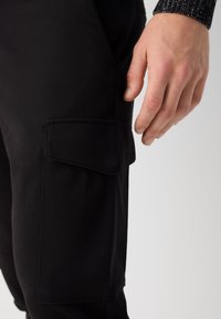 Černé nákladní kalhoty s boční kapsou, vyrobené z odolné bavlněné látky. Textura je hladká, s čistým šitím a jednoduchým designem.