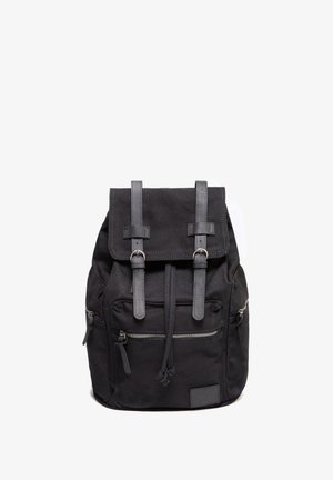 Mochila de lona negra con solapa superior y dos correas, bolsillos frontales con cremallera y bolsillo deslizante, y un panel de cuero sintético texturizado.