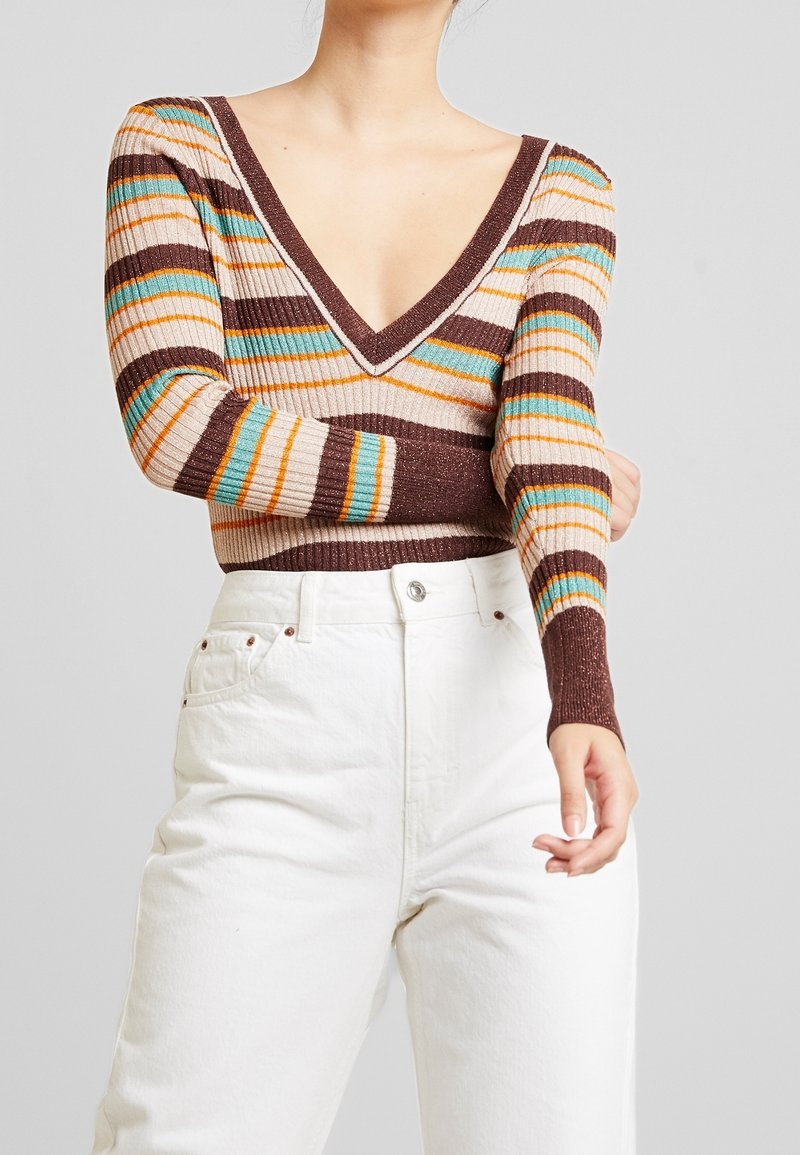 Person klädd i en djup V-ringad, ribbad tröja med bruna, rosa, teal och orange ränder och högmidjade vita jeans mot en enfärgad bakgrund.