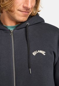 Navy zip-up hoodie featuring a soft fleece interior, black drawstring, and cream embroidered logo on the chest. Durable zipper and pockets.