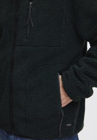 Dark green fleece jacket with a textured, soft surface, front zip closure, and a zippered pocket on the side. Features a relaxed fit.