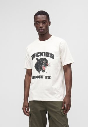 Άντρας που φοράει λευκό t-shirt με γραφικό μαύρης πάνθηρας και κείμενο "Dickies Since '22" συνδυασμένο με ελιά cargo παντελόνια σε απλό φόντο.