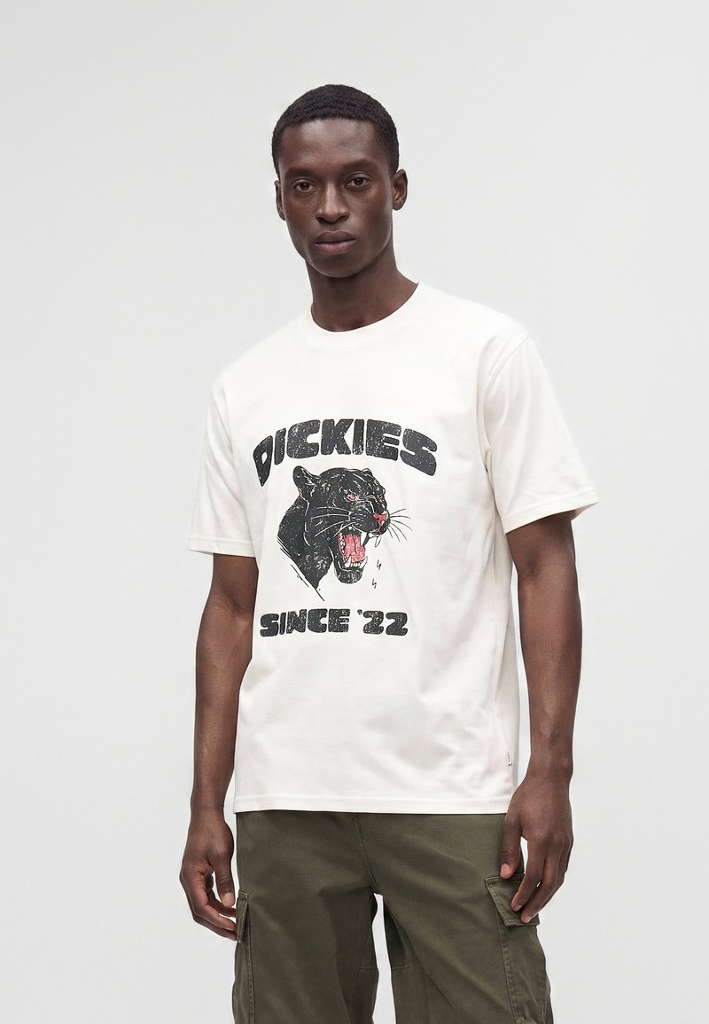 Άντρας που φοράει λευκό t-shirt με γραφικό μαύρης πάνθηρας και κείμενο "Dickies Since '22" συνδυασμένο με ελιά cargo παντελόνια σε απλό φόντο.