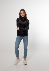 Giacca in pile nera con una texture morbida e motivo leopardato; presenta un colletto alto e una zip. Abbinata a jeans azzurri e sneakers bianche.