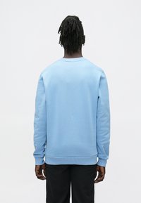 Sötét, fonott hajjal rendelkező személy világoskék hosszú ujjú pulóvert és fekete nadrágot visel, háttal áll egy egyszerű fehér háttér előtt.