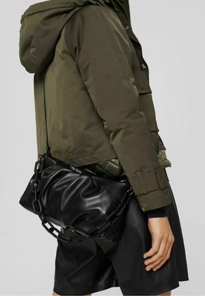 Persoon draagt olijfgroen jack en zwarte leren rok, met een zwarte, geplooide schoudertas met een dikke kettingband.
