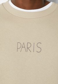 Beige sweatshirt met geribbelde ronde hals; "PARIS" gedrukt in een gedempte bruine lettertype in het midden. Zachte textuur, casual design.