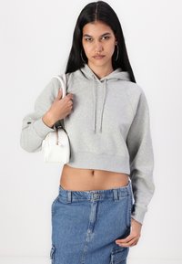 Harmaa cropped-huppari, jossa on nyörit, yhdistettynä farkkuhameeseen. Malli pitää pientä valkoista käsilaukkua. Pitkät tummat hiukset, renkulakorviksia.