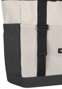 Taška so zmesou béžovej tkaniny a čiernych akcentov, s textúrovaným sieťovaným panelom, pevným popruhom a obdĺžnikovým tvarom.
