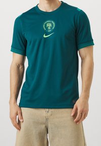 Camiseta deportiva azul verdoso hecha de tela que absorbe la humedad, con cuello redondo, mangas cortas, logotipo de la Federación de Fútbol de Nigeria y rayas.