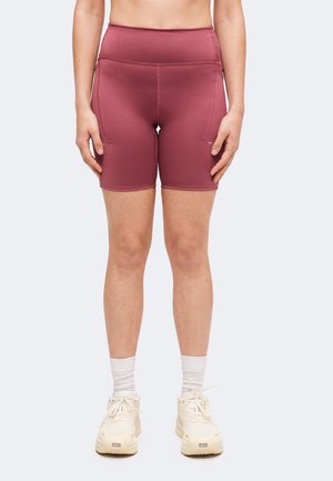 Person, der har højtaljede mauve cykelshorts, hvide sokker og hvide Nike sneakers, står mod en ensartet lys baggrund.
