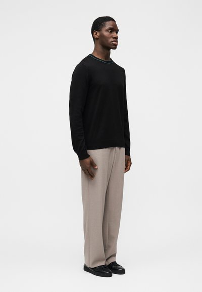 Čierny sveter s dlhým rukávom a zaobleným výstrihom so zeleným lemovaním, v kombinácii s svetlobéžovými širokými nohavicami a čiernymi topánkami.