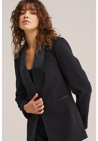 Kvinna med lockigt brunt hår som bär en svart kavaj med sidenuppslag, håller framkanten mot en neutral bakgrund.