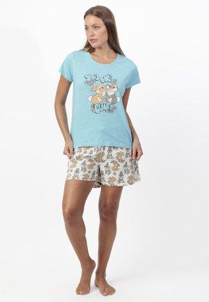 Mujer de pie con camiseta azul y pantalones cortos blancos con dibujos de conejos de dibujos animados y texto «¿Estás bien, culo?»