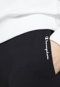 Svarta bomullsbyxor har en tight passform med en vit broderad "Champion"-logotyp på sidan och en mjuk textur.