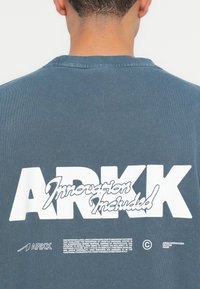 Tricou scurt cu mâneci, de culoare albastru închis, cu text alb imprimat pe spate, inclusiv logo-ul "ARKK" și mesajul "Inovație inclusă".