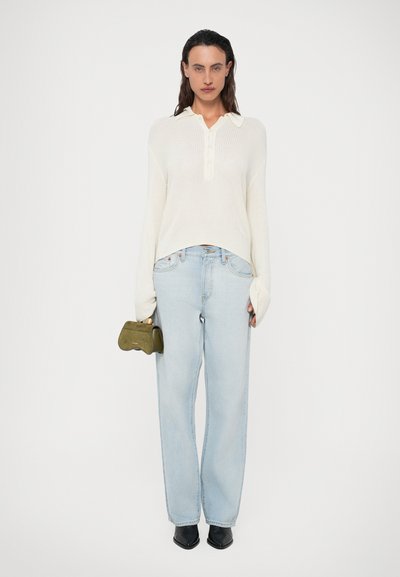 Mulher vestindo um suéter de malha creme e jeans azul claro, segurando uma pequena bolsa verde oliva, de pé contra um fundo branco simples.