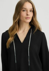 Zwarte top met een V-hals en lange mouwen, voorzien van een strook ronde metalen accenten aan de voorkant. Glad materiaal en een losse pasvorm.