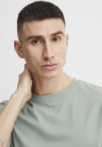 Világoszöld, rövid ujjú póló kerek nyakkal. A modell keze a nyakán pihen, ezzel kiemelve a sima anyagot és a letisztult dizájnt.