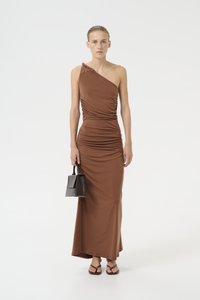 Brown one-shoulder dress with gathered detailing, floor-length design, and a smooth texture. Model holds a small black handbag.