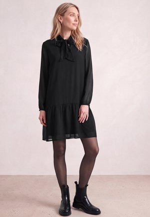 Une femme porte une robe noire à manches longues avec un col à nœud, des collants transparents et des bottines, devant un fond de studio clair.