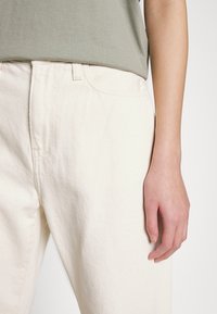 Kermanväriset puuvillahousut, joissa on tekstuurillinen kangas, esillä yhdessä käden kanssa, joka on pukeutunut vaaleanvihreään yläosaan, ja housujen rento istuvuus on nähtävissä.