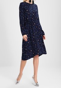 Navyblaues Kleid mit langen Ärmeln, gerafftem Taillengürtel und Herz- sowie Kreuzmustern in helleren Farben. Getragen mit metallischen High Heels.