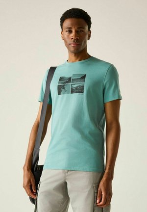 Hombre vestido con camiseta gráfica verde azulado y pantalones cortos cargo beige lleva un bolso negro al hombro sobre fondo liso.
