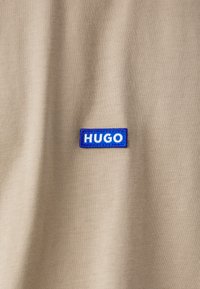 Beige stof t-shirt med en blå rektangulær mærkat med hvid "HUGO" tekst syet på forsiden. Glat tekstur, afslappet design.