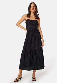 Abito maxi senza spalline di colore nero con corpino in pizzo e gonna a strati. Presenta una vita aderente e un design fluido, realizzato in tessuto leggero.