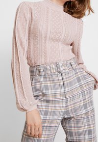 Jasnoróżowa, dziana bluzka z teksturowymi wzorami i długimi rękawami, zestawiona z wysokopasa spodniami w kratę w wielokolorowy wzór.