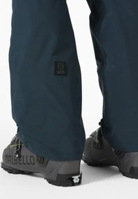 Marine outdoorbroek met een textuurstof en een logopatch op de dij, gecombineerd met grijze snowboardlaarzen met verstelbare hardware en zwarte zolen.