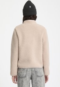 Beige fleecejack met een zachte textuur, hoge kraag en lange mouwen. Gecombineerd met lichtgrijze, vervaagde denim broek en een zwarte gebreide beanie.