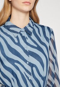 Hele sinine särgike tumeda sinise sebratriibulise mustriga, pehme tekstuuriga ja klassikalise kraega, millel on väikesed nööbid.